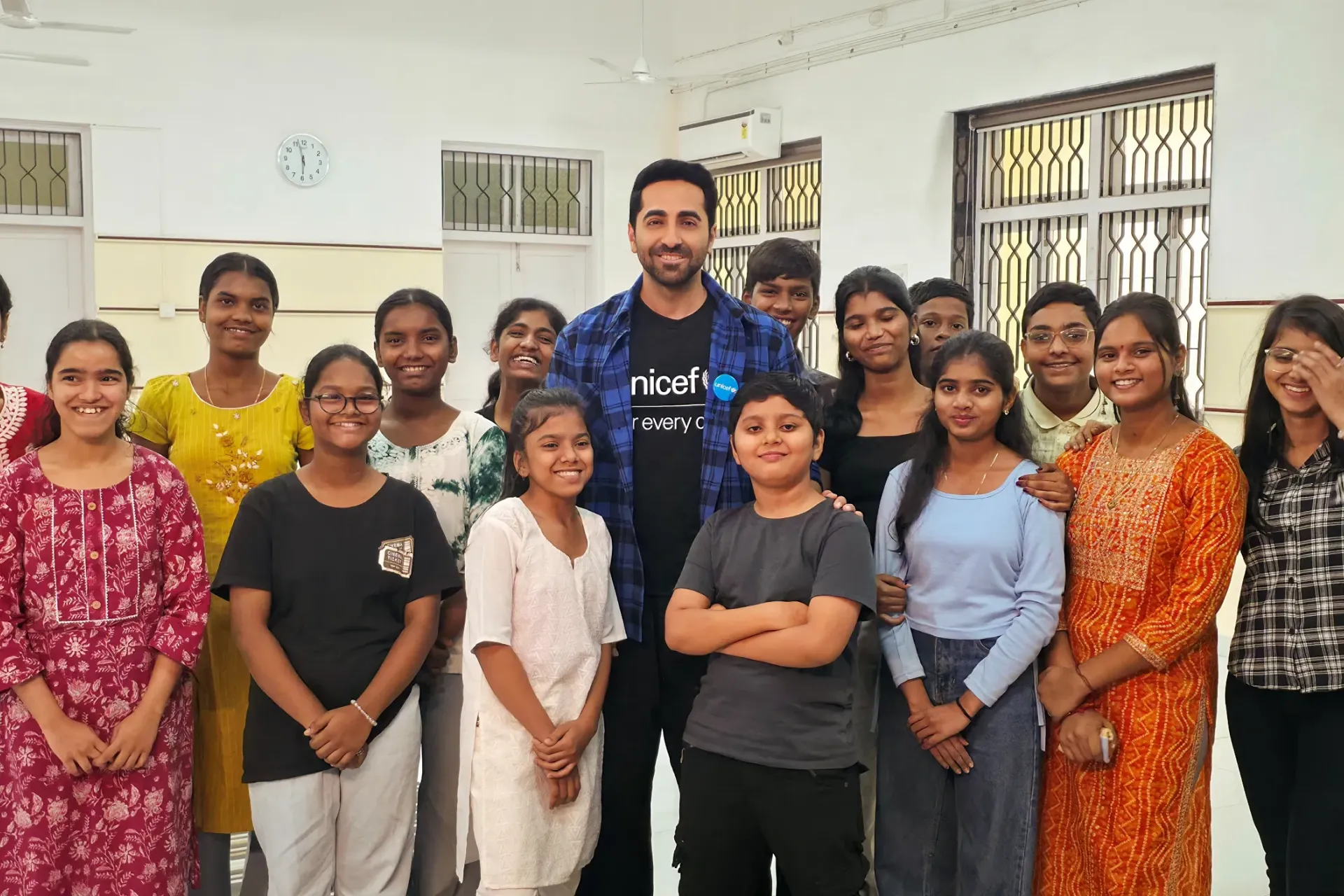UNICEF India National Ambassador Ayushmann Khurrana poses warmly with a group of smiling children and adolescents in a community centre, wearing his UNICEF "for every child" t-shirt.