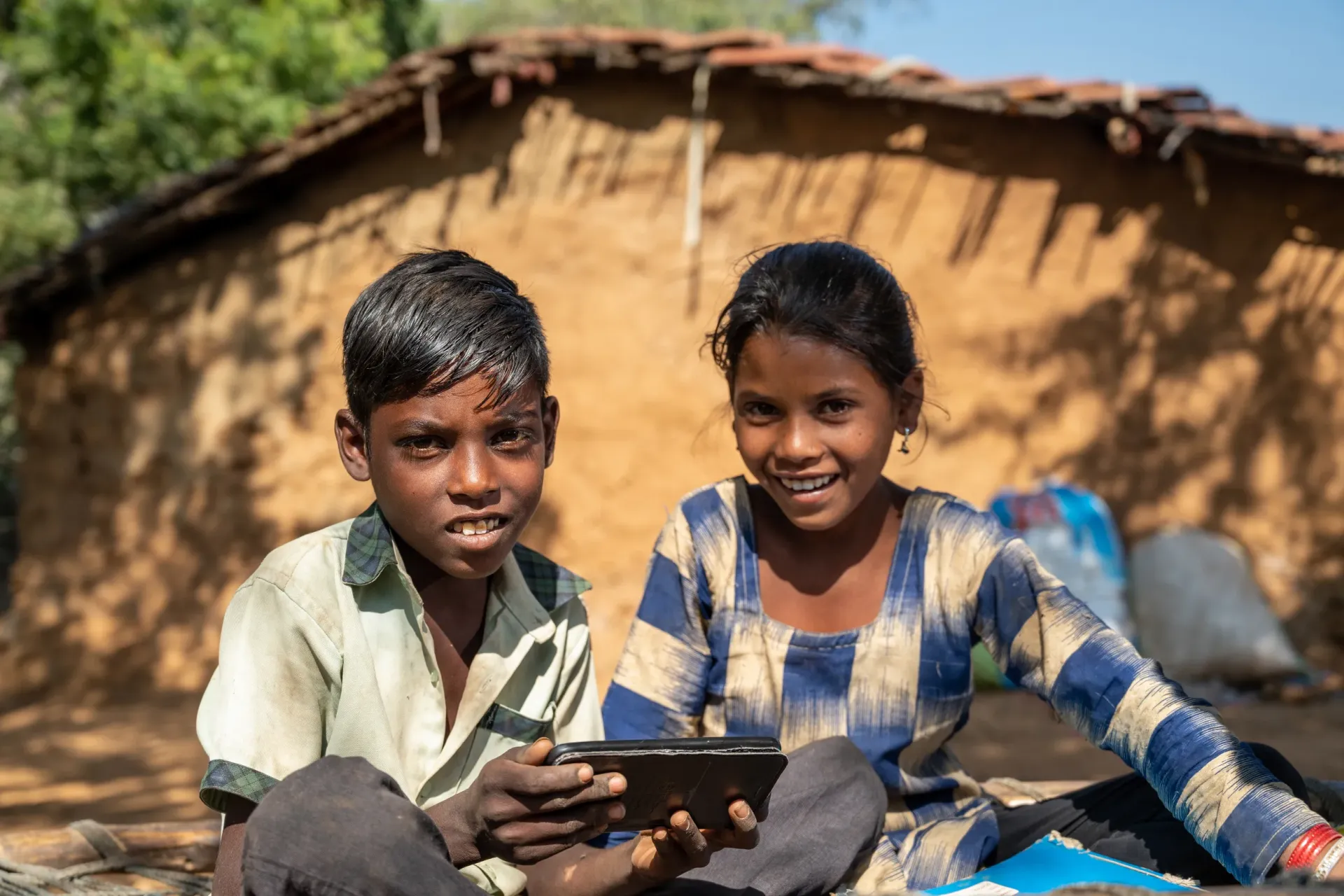A boy and a girl sit together outside a mud-walled village home, sharing a tablet device, as the girl smiles warmly at the camera.