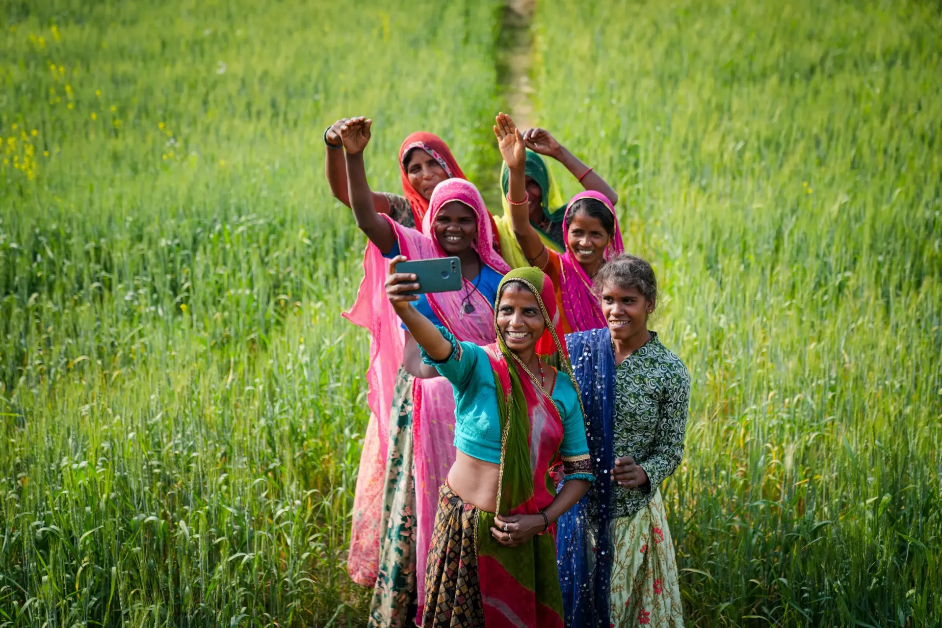 A group of joyful Indian women and girls in colourful sarees take a selfie together in a lush green crop field, waving and smiling broadly.