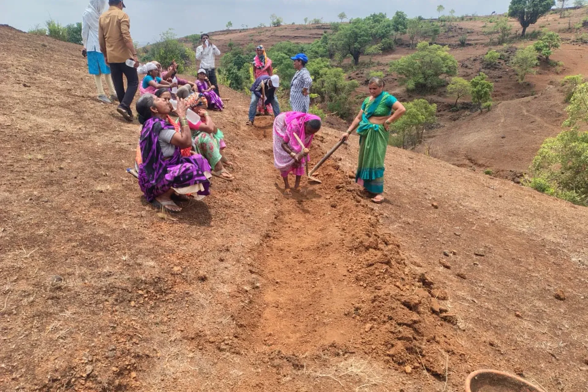 A group of rural women in colourful sarees dig and hoe a Continuous Contour Trench on a dry red-soil hillside in Maharashtra during a community Shramdaan water conservation campaign.
