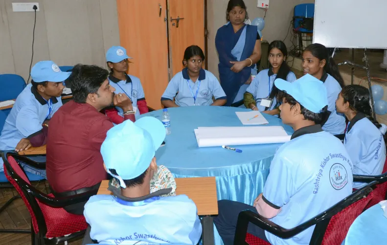Tribal adolescents in RKSK uniforms sit in a group discussion around a round table with a facilitator, as part of a mental health stigma interaction session in Koraput, Odisha.