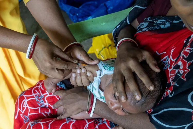 A health worker administers an injectable vaccine into the arm of two-month-old Pallavi Roy during a routine immunization outreach session. The baby lies cradled across her mother's lap, dressed in a white outfit, while the mother gently steadies her head and holds her arm still. The women's colourful sarees and glass bangles frame the scene from above.