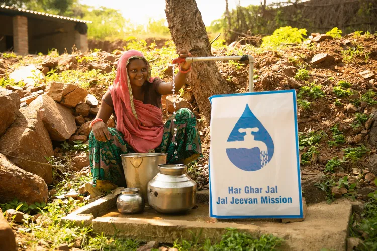 A smiling woman in a pink dupatta turns on a community water tap beside a 'Har Ghar Jal &mdash; Jal Jeevan Mission' sign, filling steel vessels in a rural setting.
