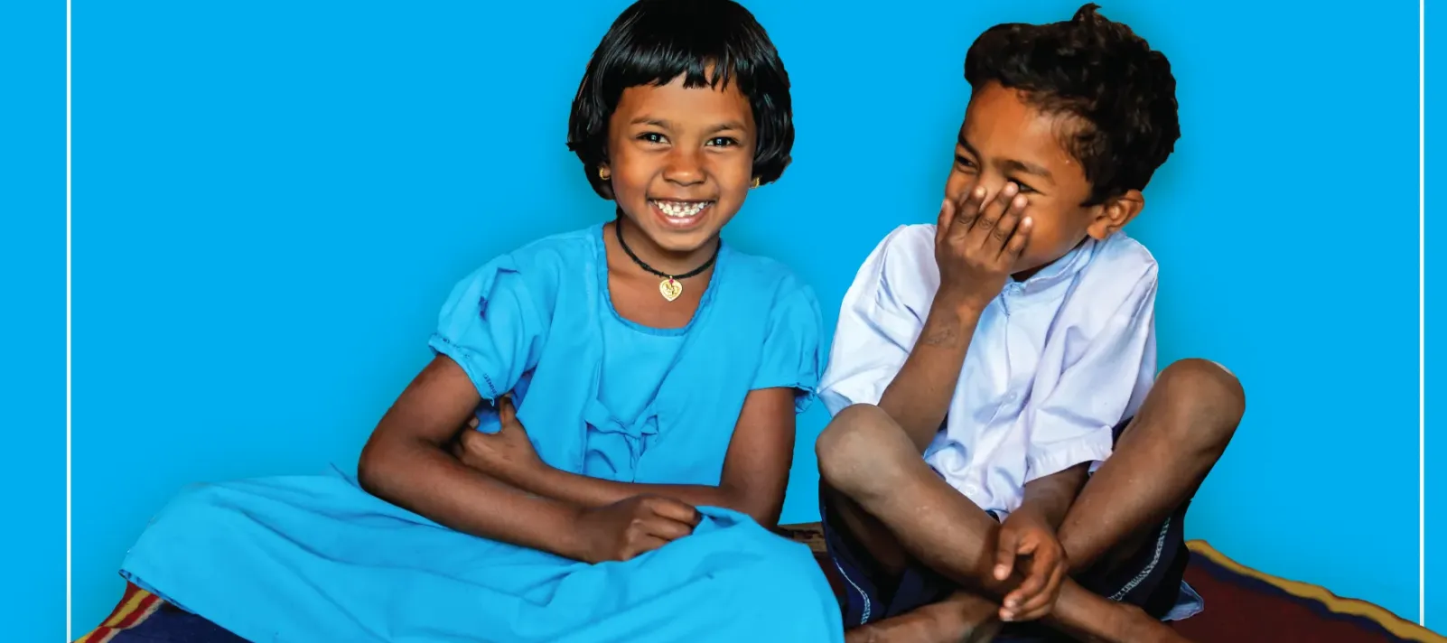 Two joyful young Indian children &mdash; a smiling girl in a blue dress and a giggling boy in white &mdash; sit together on a colourful mat against a bright blue background, representing childhood happiness, child rights, and UNICEF India's 2023 Calendar.