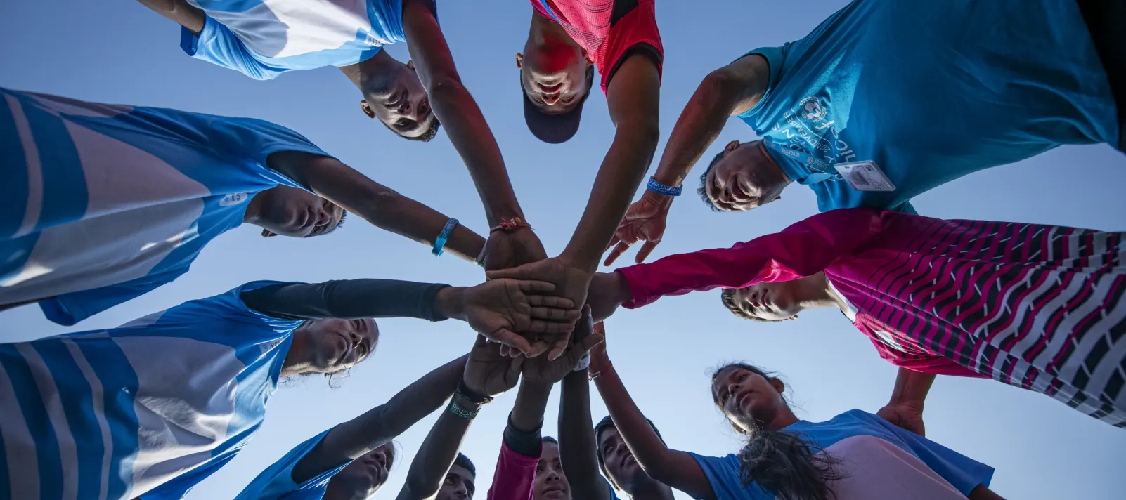 A low-angle shot looking up at a circle of young girls in blue and pink sports jerseys stacking their hands together in a team huddle against a clear blue sky.