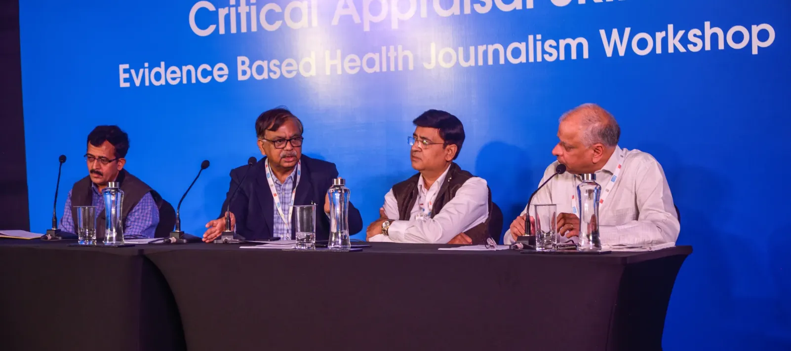 Four male panellists sit at a conference table during the "Critical Appraisal Skills &mdash; Evidence Based Health Journalism Workshop" organised by MANUU, as indicated by the blue backdrop behind them.
