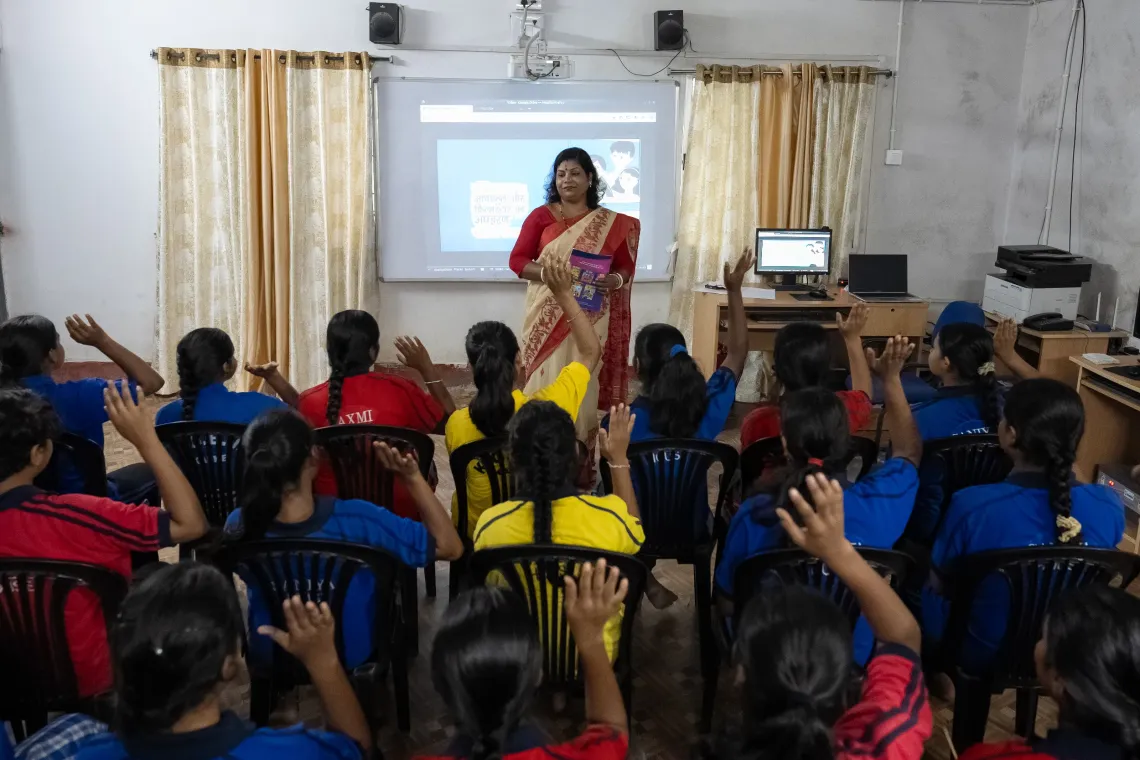 A teacher in a saree leads an interactive digital classroom session on adolescent health, as a room full of girls in blue uniforms eagerly raise their hands.
