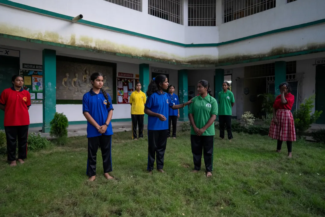 A group of Indian schoolgirls in colourful house uniforms stand barefoot on a school courtyard lawn during what appears to be an outdoor activity or role-play session.