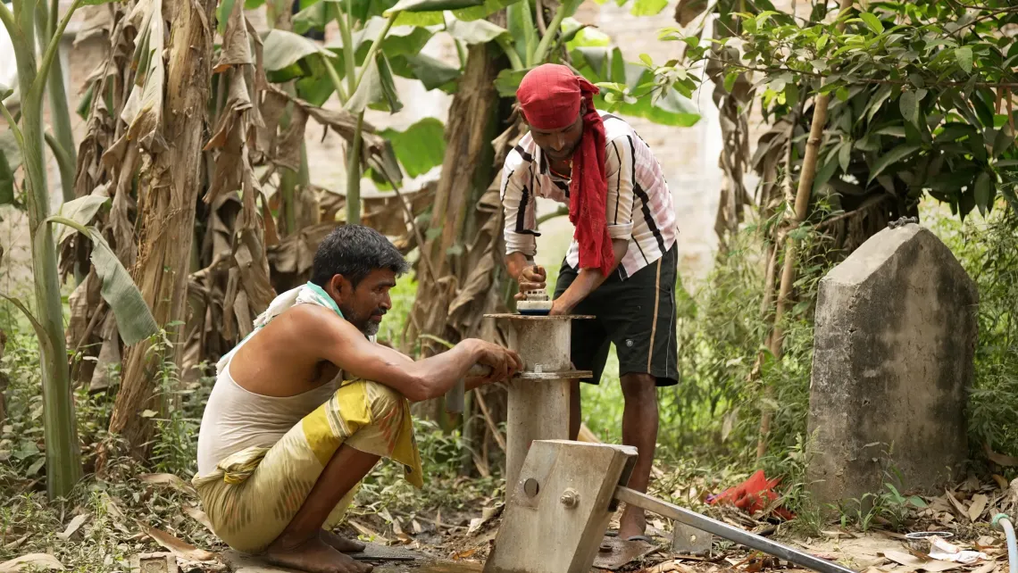 Two men work together to install a water pump or standpipe fitting in a lush rural setting surrounded by banana trees and vegetation.