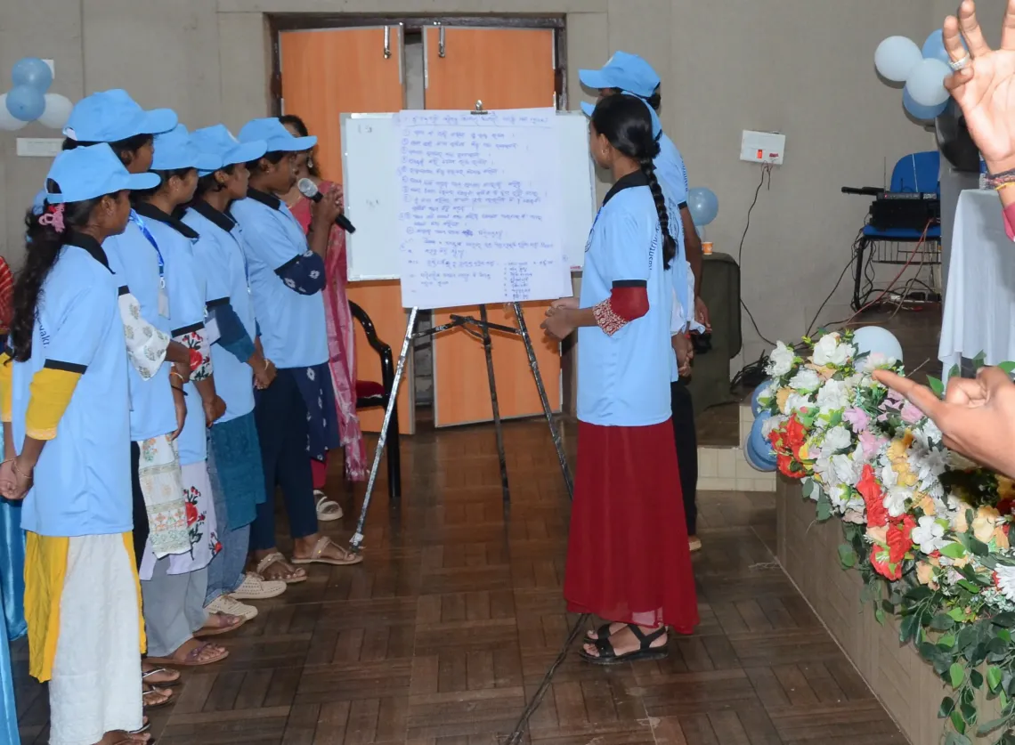 A tribal adolescent girl presents her group's findings on mental health stigma at a whiteboard covered in Odia notes, as fellow RKSK-uniformed peers stand listening behind her in Koraput, Odisha."