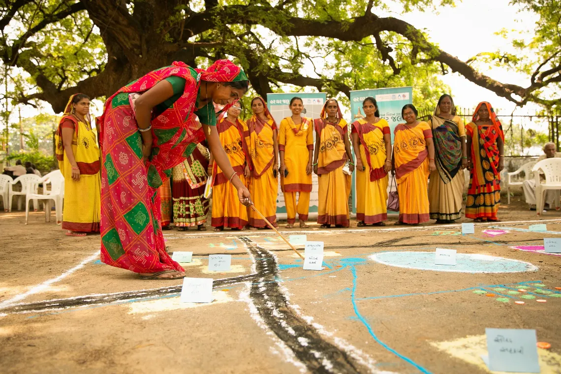 Women in yellow saris watch as a woman in red sari creates rangoli outdoors beneath a large tree.