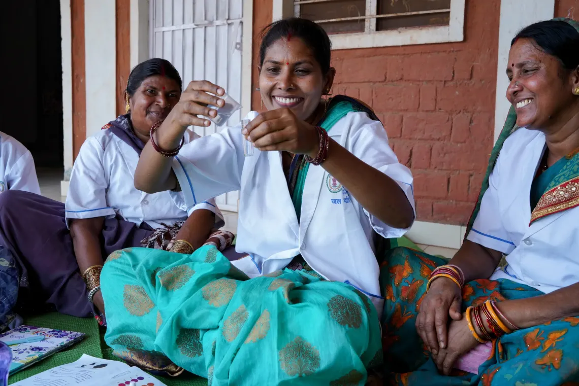 Smiling woman in a white coat holds up a small cup, seated with two others by a brick wall.