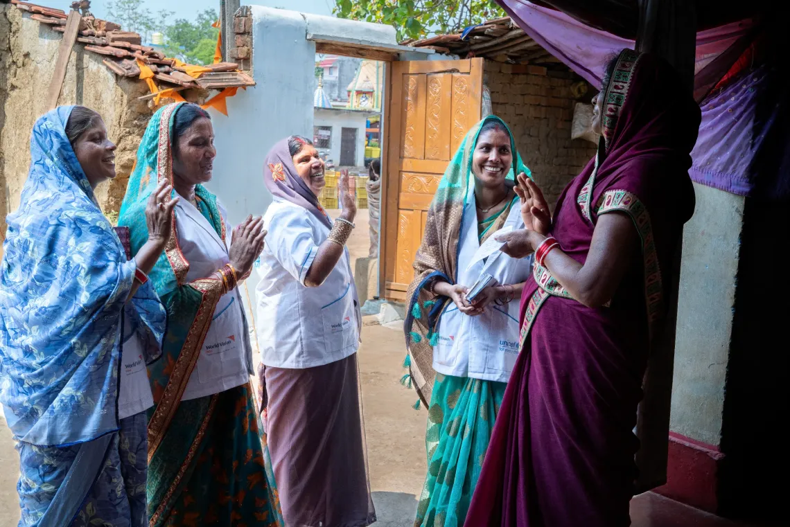 A group of smiling Indian women in colourful sarees and World Vision-UNICEF white coats laugh and interact warmly during a community home visit in a rural village lane.