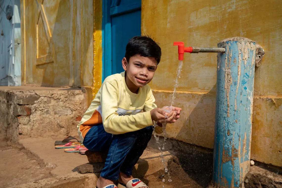 A young Indian boy in a yellow sweatshirt crouches beside a community water tap, washing his hands with clean running water outside a yellow-walled village home.