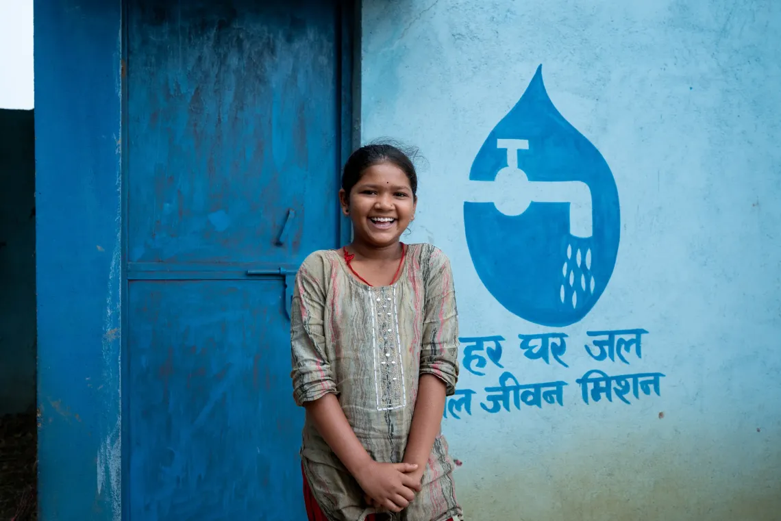 A smiling Indian girl stands beside a blue wall painted with the Jal Jeevan Mission logo and the Hindi slogan "Har Ghar Jal" &mdash; clean water for every home.