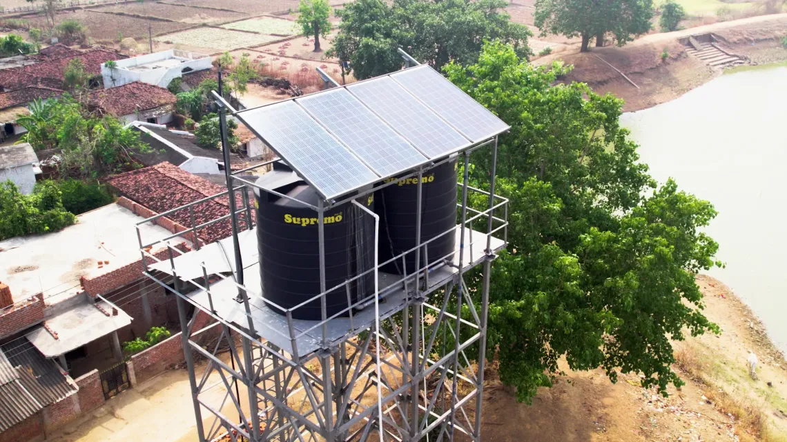 An aerial view of a solar-powered water tower with three black "Supremo" storage tanks and solar panels mounted on a metal frame, serving a rural Indian village beside a river.