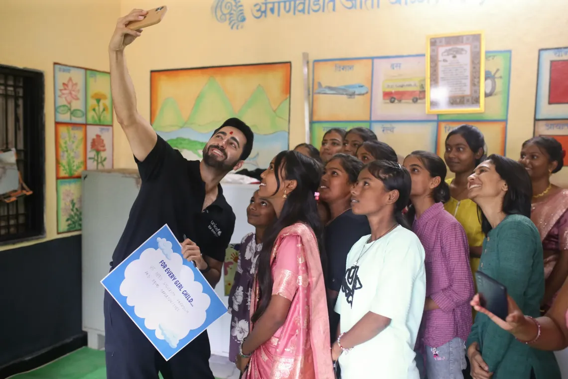 Ayushmann Khurrana, UNICEF India National Ambassador, in a black UNICEF polo shirt and a red tilak on his forehead, holds up his phone to take a selfie with a group of laughing and excited adolescent girls inside a colourfully decorated Anganwadi centre in Maharashtra.
