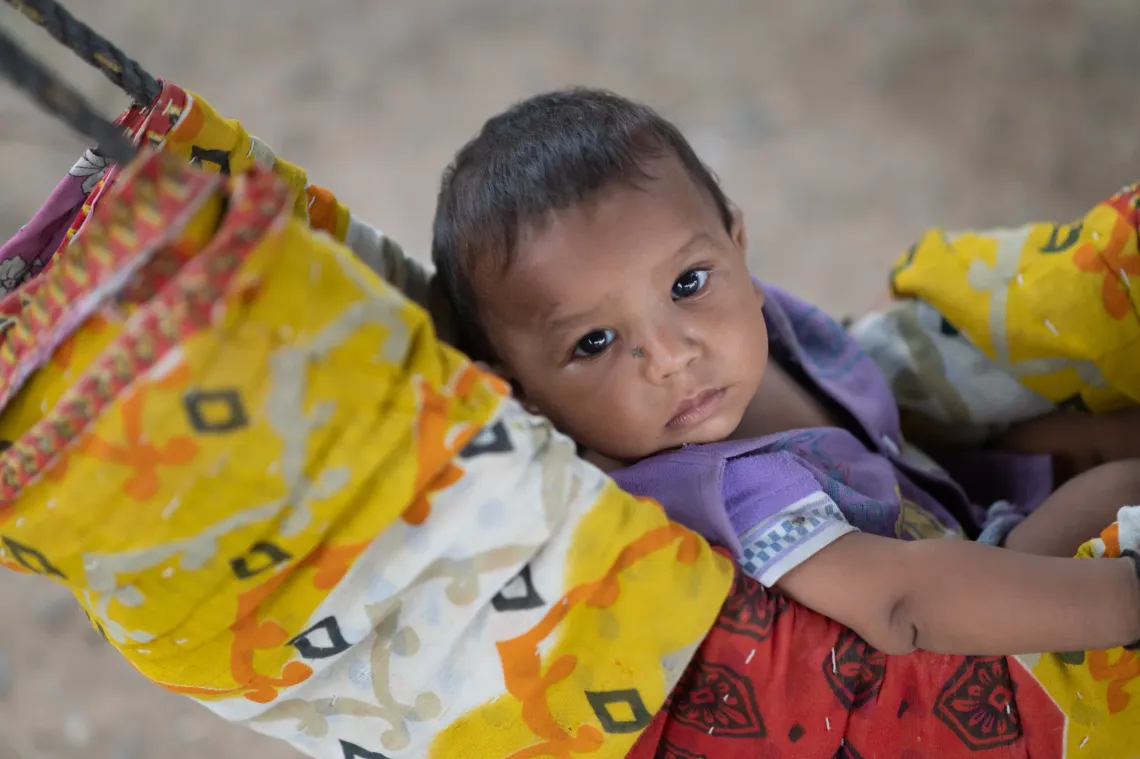A young Indian baby with large expressive eyes rests in a colourful fabric hammock cradle, gazing quietly at the camera.