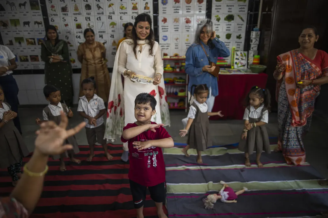 A young boy dances joyfully at the centre of an early childhood learning space, while a woman in a white floral kurta, other adults and toddlers in school uniforms look on smiling. Gujarati alphabet charts and colourful toys fill the background.