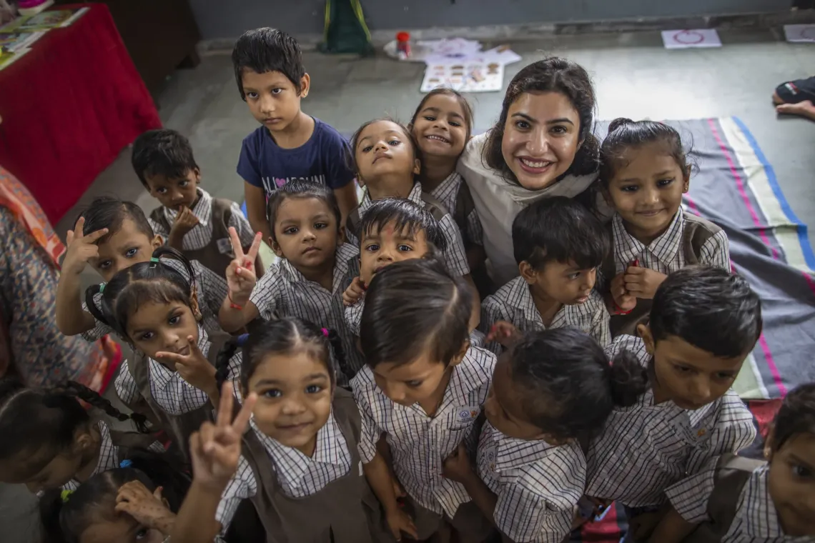 A smiling woman is surrounded by a joyful group of young children in brown striped school uniforms in a classroom, several flashing victory signs at the camera.