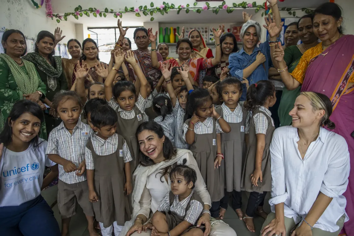 A laughing woman in white sits at the centre of a joyful group photo with young children in school uniforms, community women and UNICEF staff inside a decorated early childhood centre.