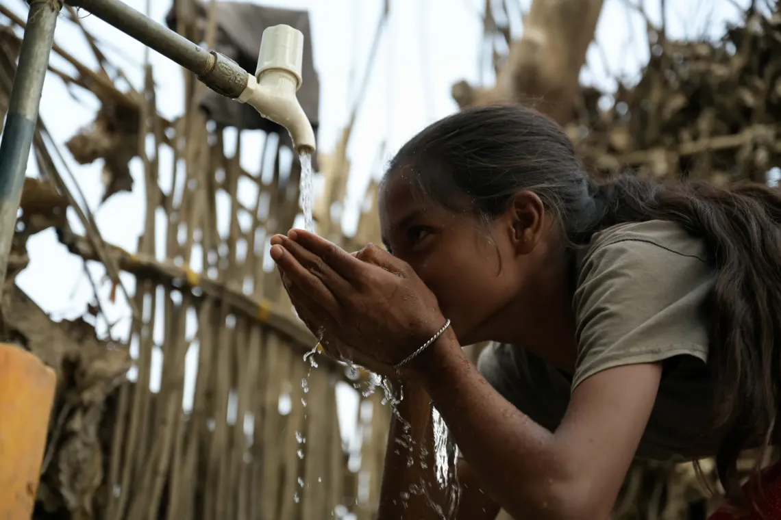 Child cupping hands to drink from outdoor tap in a rustic setting.