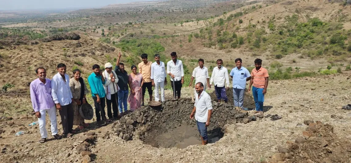 A group of community members and field staff stand together on a dry hillside beside a freshly dug water harvesting trench, with a vast drought-prone landscape stretching behind them in Maharashtra.
