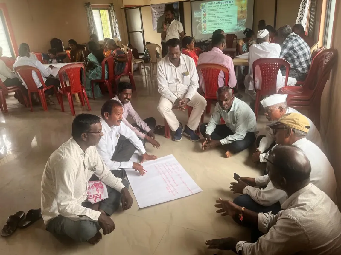 Community men including farmers and Gram Panchayat members sit in small groups on the floor and chairs during a UNICEF-WOTR climate resilience workshop in Maharashtra, reviewing Marathi-language planning charts while a climate presentation is projected on screen.