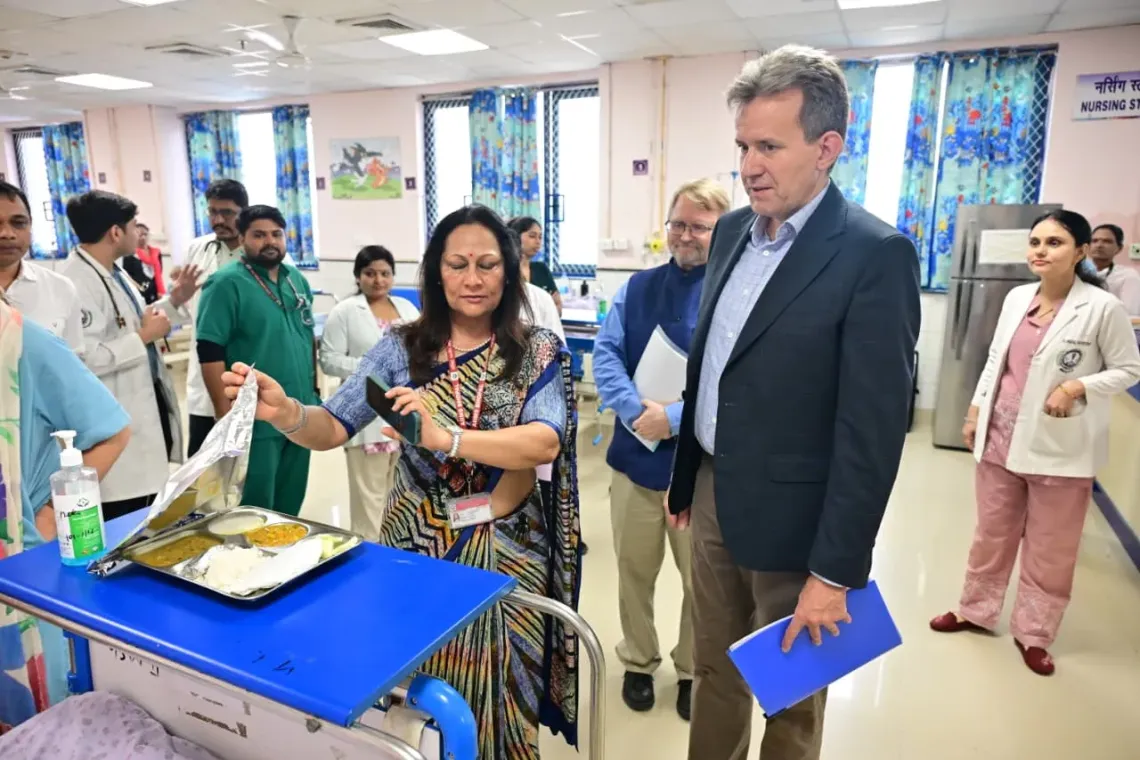 Jesper Moller, Deputy Representative &ndash; Programmes, UNICEF India, watches as a senior doctor in a saree lifts the foil cover from a meal tray with rice, dal, vegetables, and curd in a pediatric ward, surrounded by doctors, nurses, and medical staff in white coats and scrubs, with children's ward decorations in the background.