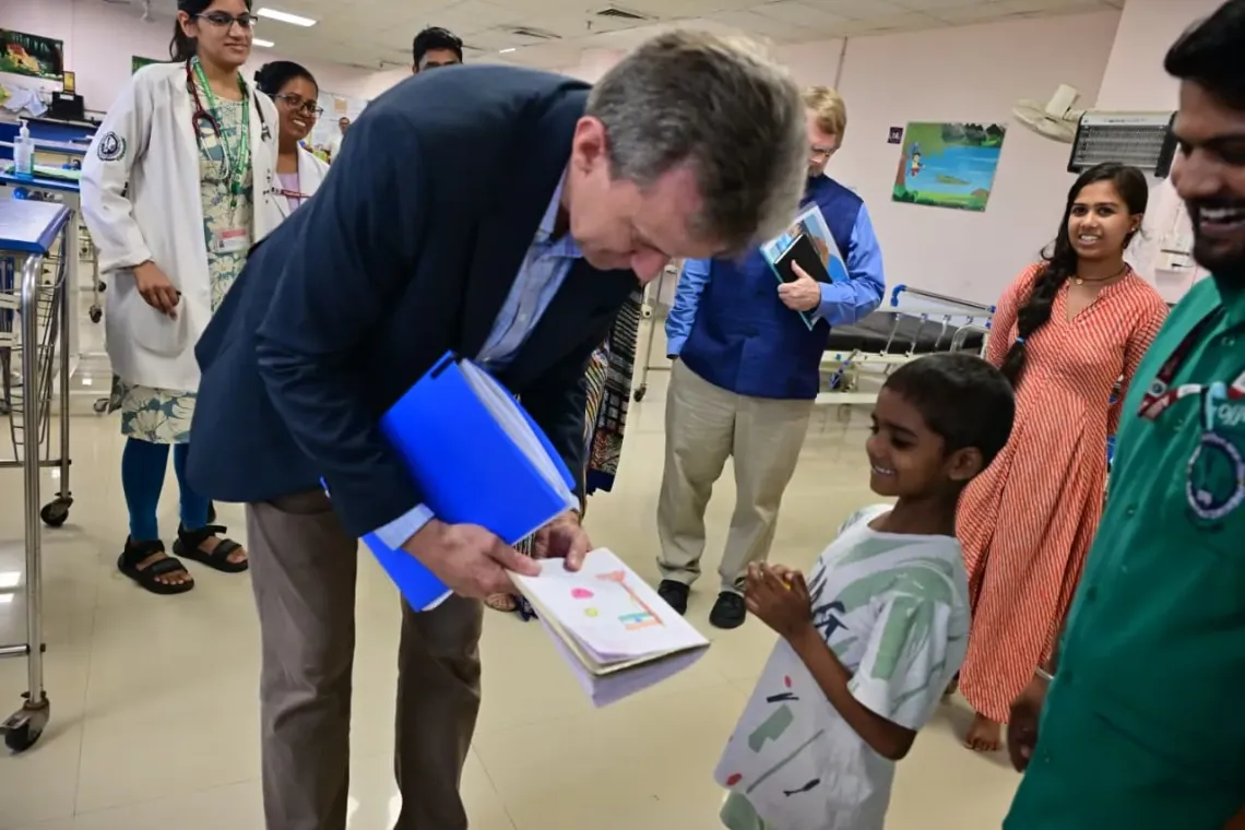 Jesper Moller, the Deputy Representative for Programmes at UNICEF India, bends down with a warm smile to admire a drawing presented to him by a cheerful young boy in a pediatric ward. The child's mother and hospital staff, including doctors in white coats, watch happily in the background.