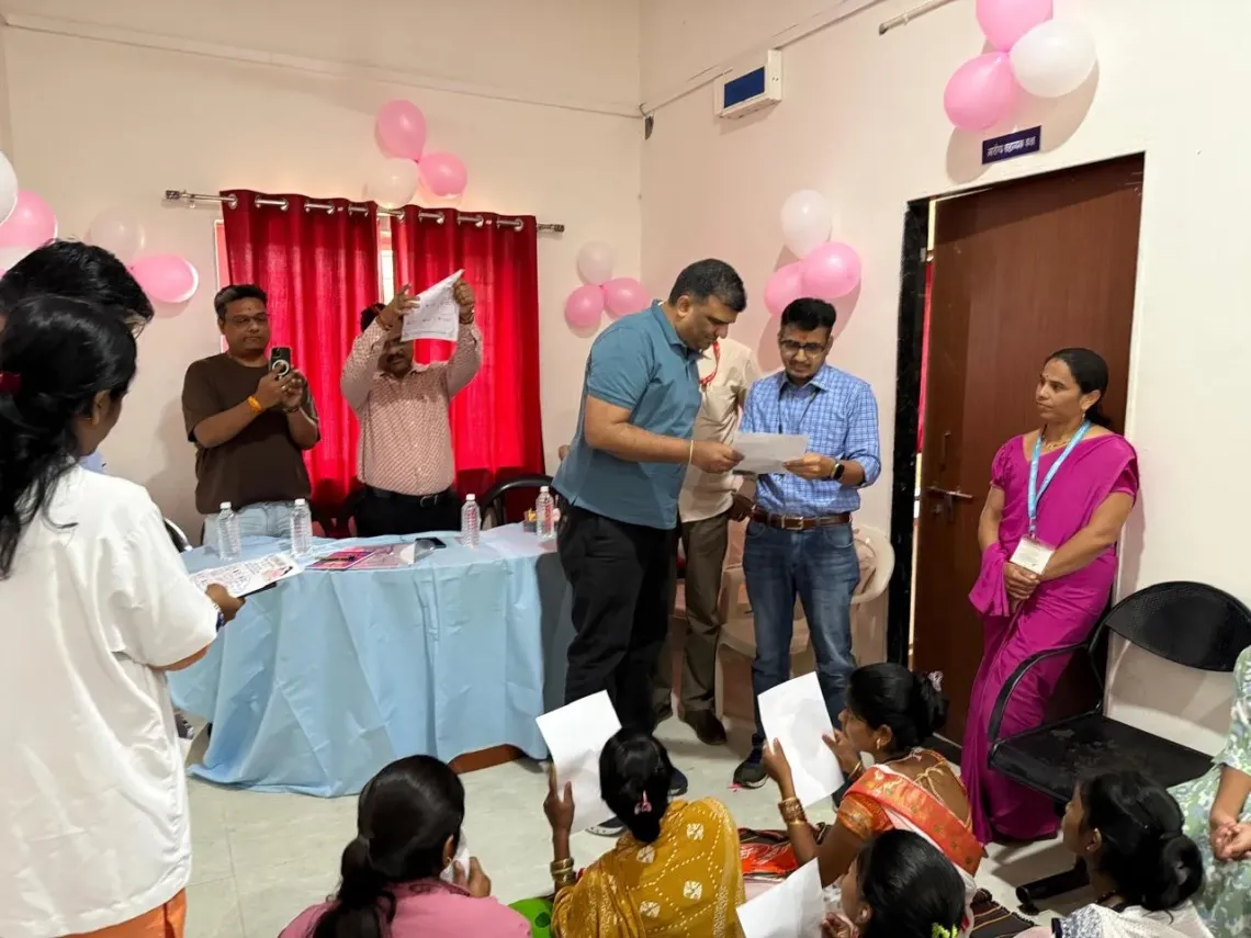Women seated on the floor hold up sheets of paper while visitors and health staff review materials in a balloon-decorated room at a health centre