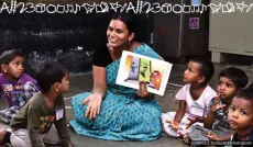An Anganwadi worker in a blue saree reads a colourful picture book to a group of attentive young children sitting around her on the floor.