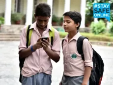 Two Indian schoolboys in pink uniforms with backpacks look at a smartphone together in a school courtyard, with the #StaySafeOnline hashtag in the top right corner.
