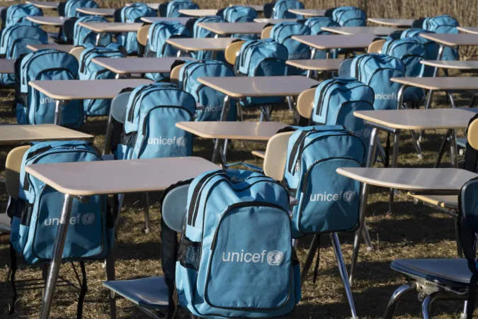 Blue backpacks with UNICEF logos hand on empty school chairs and desks placed on grass outside.