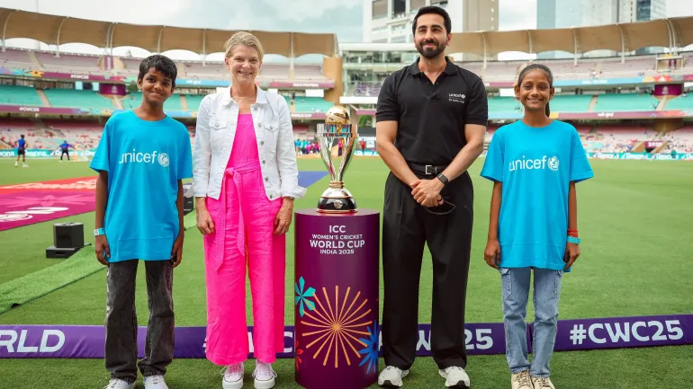 Ayushmann Khurrana is joined by Cynthia McCaffrey and a boy and girl while they stand beside the ICC Women's Cricket World Cup Trophy in a stadium
