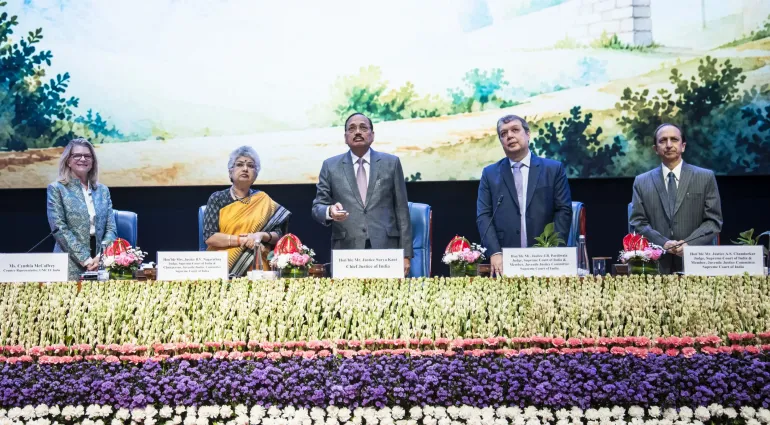 Three officials on stage behind lush floral display; central figure speaks into microphone before scenic backdrop.
