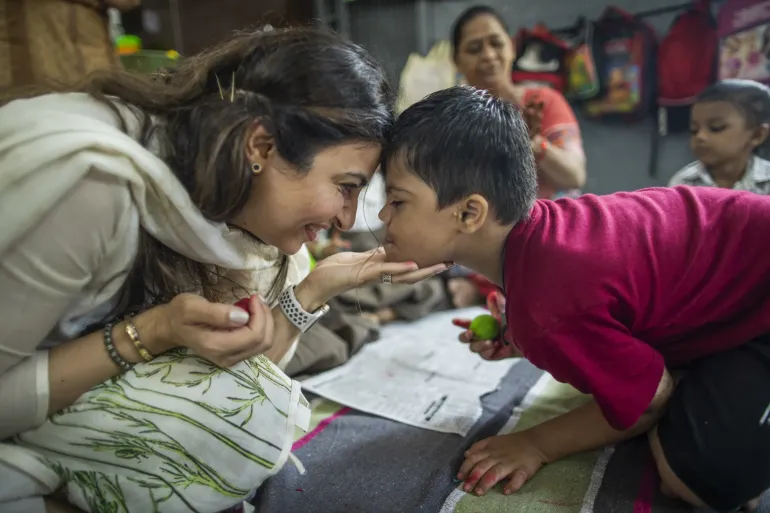 A woman and a young boy touch foreheads and laugh together in a warm, playful moment on the floor of a classroom or community learning space.