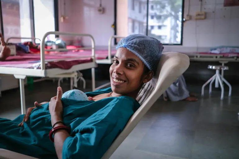 A smiling new mother in a hospital gown and surgical cap holds her newborn baby while reclining in a chair in a maternity ward.