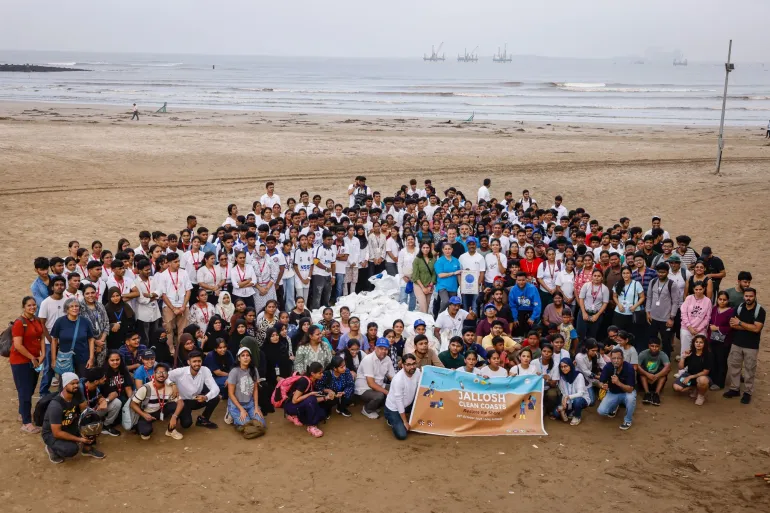 A large group of diverse young people, including students and volunteers wearing casual clothes and some with red lanyards, pose for a group photo on a sandy beach. They are arranged in multiple rows, standing, sitting, and kneeling, forming a semi-circle. In the front, a few individuals hold a banner reading "JALLOSH CLEAN COASTS Restore the Shore" with the date "29th October 2023 | Juhu, Koliwada" and icons of people cleaning. The background shows the ocean with gentle waves.