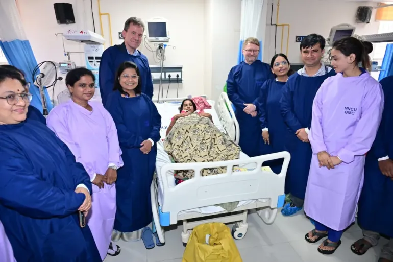 UNICEF officials and hospital staff in navy blue and lavender gowns gather around a mother resting under a blanket in the Mother and Newborn Care Unit. Jesper Moller, UNICEF India's Deputy Representative for Programs, is present, with medical equipment visible in the background.