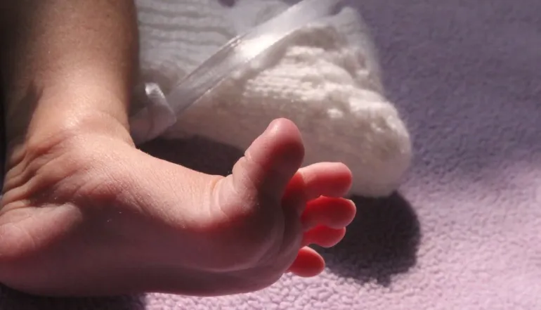 A close-up of a baby's bare foot resting on a soft lilac blanket, lit by warm sunlight that highlights the tiny toes and delicate skin. A white knitted bootie lies just behind the foot.