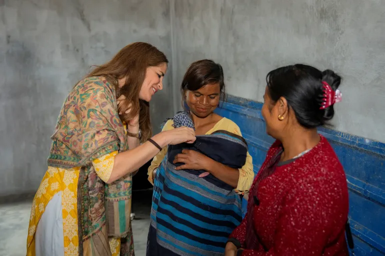 UNICEF India's Chief of Field Services leans in warmly to look at a newborn wrapped in a striped cloth, held by a young mother, as a Buddy Mother looks on. The interaction takes place against a grey wall with a blue border, reflecting a grassroots community health visit in rural Meghalaya.