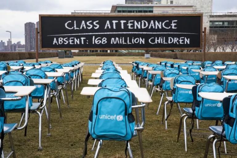 Blue UNICEF backpacks sit on school chairs and desks set out in a park on grass. On a blackboard there are the words 'class attendance, absent 168 million children'.