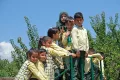 A group of young schoolchildren in yellow checked uniforms cluster together on outdoor playground equipment under a bright blue sky, with one girl wearing a green hijab standing behind them.