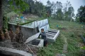 A man inspects a rural water filtration and storage system, consisting of concrete tanks and a gravel filter bed, on a hillside surrounded by trees and farmland.