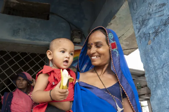 A smiling mother in a blue saree holds her toddler in red who eagerly reaches for a banana outside a community setting.