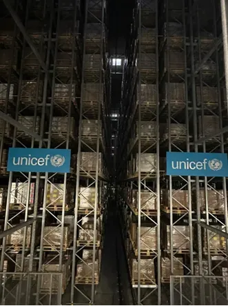 Tall metal shelving racks stacked with palletised supplies stretch down a long aisle in a UNICEF warehouse, with blue UNICEF signs mounted on either side.