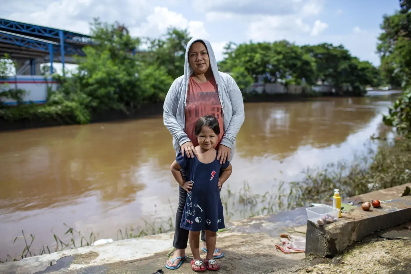 A girl and her mother stand in front of a river that overflowed and inundated their home