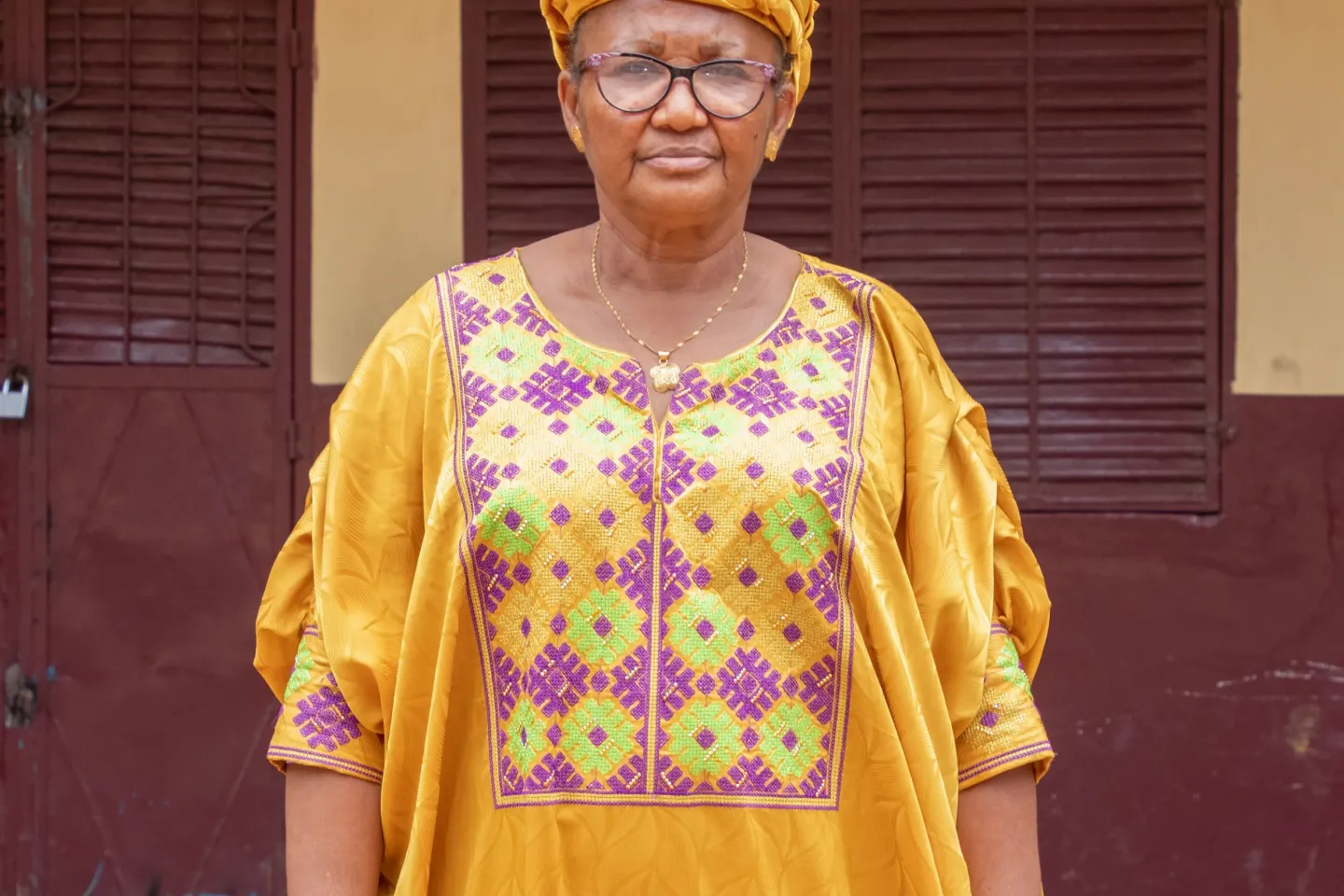 A woman, a teacher, poses in front of a school and stares at the camera.