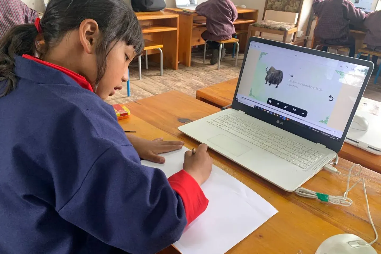 A girl writes on a paper with a laptop in front of her in a school classroom.